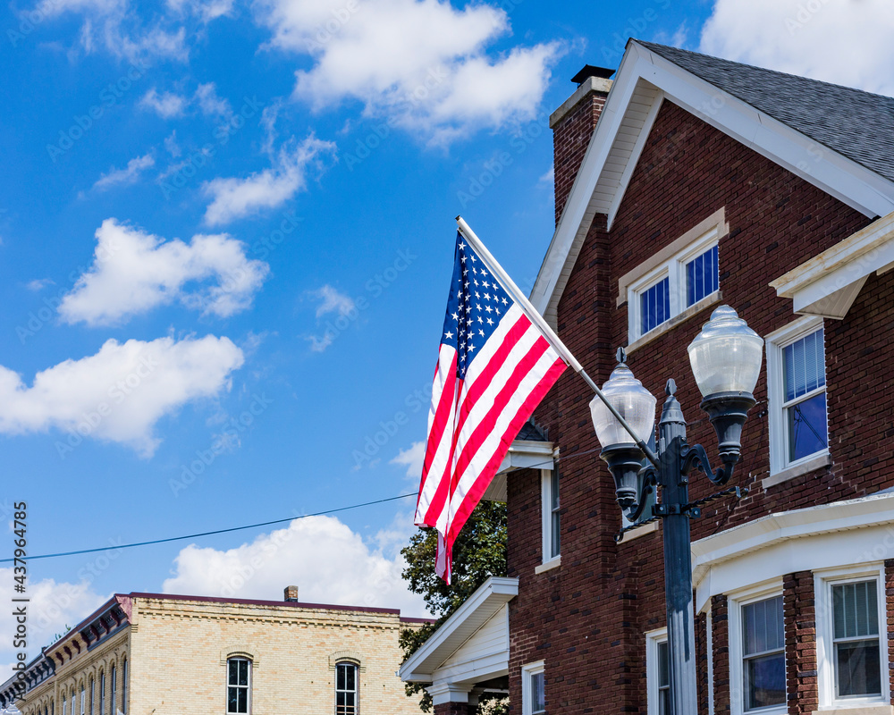 An American flag on a light pole with two vintage street lights and an old, brick building in the background with a blue sky and white clouds.