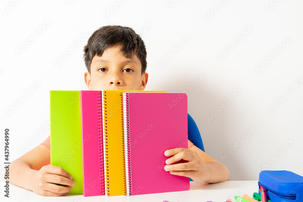 Boy showing various colored school notebooks sitting on desk. Various colored pencils on desk next to his pencil case and other school supplies. Back to school concept. Boy showing various colored school notebooks sitting on desk. Various colored pencils on desk next to his pencil case and other school supplies. Back to school concept.