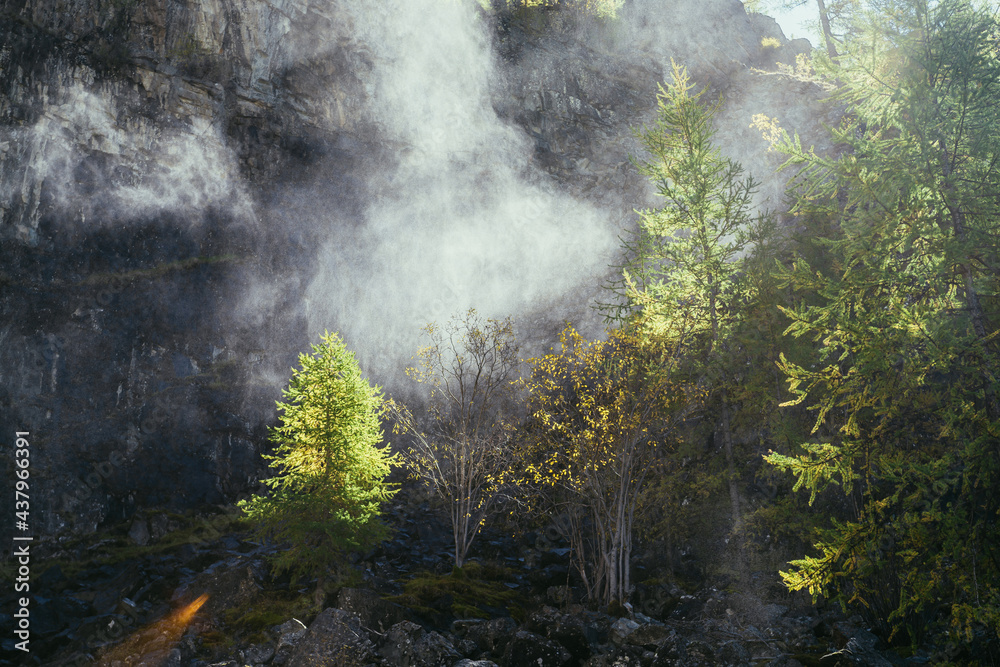 Bright landscape with water cloud of drops from waterfall on background of rocky mountain wall with autumn trees in golden sunlight. Beautiful water cloud near rock above yellow trees in gold sunshine Bright landscape with water cloud of drops from waterfall on background of rocky mountain wall with autumn trees in golden sunlight. Beautiful water cloud near rock above yellow trees in gold sunshine
