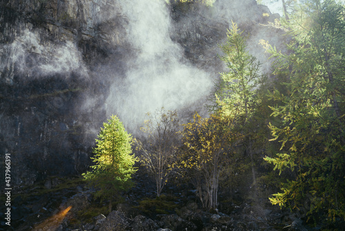 Bright landscape with water cloud of drops from waterfall on background of rocky mountain wall with autumn trees in golden sunlight. Beautiful water cloud near rock above yellow trees in gold sunshine