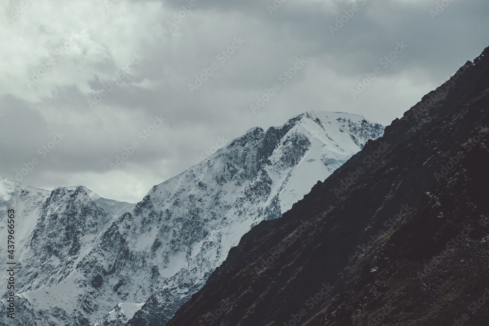 Atmospheric landscape with great snowy mountain wall in clouds. Gloomy mountain scenery with high snow-covered peaked top under cloudy sky in faded tones. Snowy pointy peak and rocks in dark colors.