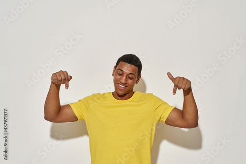 Smiling young man in casual yellow t shirt looking down and pointing with index fingers while presenting your product, standing isolated over gray background photo