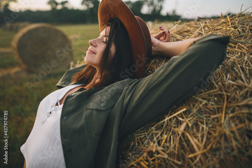 Fotografia Beautiful stylish woman in hat relaxing on haystack in summer evening field