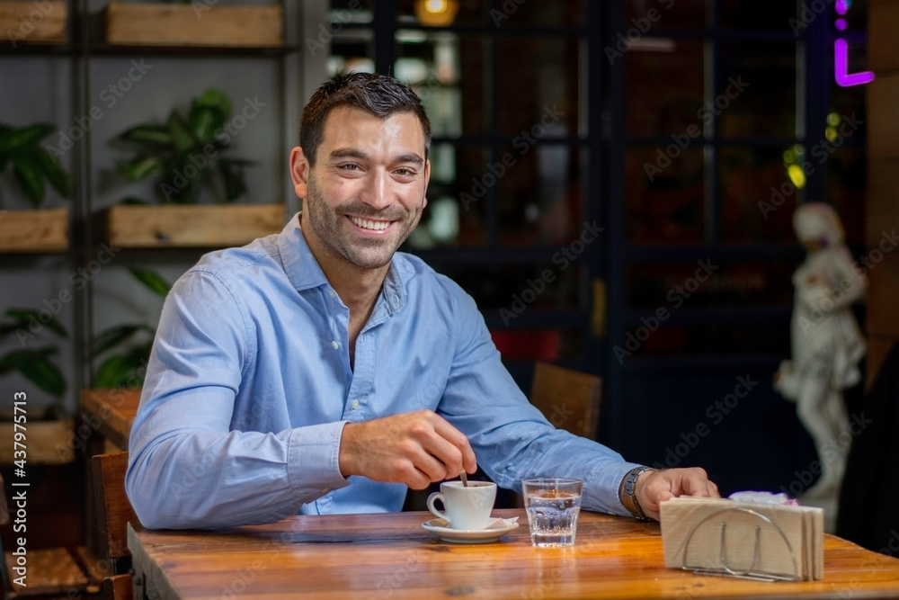 Hombre bebiendo cafe con su laptop en un descanso del trabajos sentado en una mesa de madera y una camisa celeste sonriendo a camara