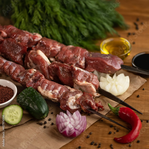 Raw marinated meat on skewers for baking. Kebab preparation and cooking Process. Sirloin, spices and vegetables on wooden table. Blurred background. Close up shot. Soft focus. Square format.