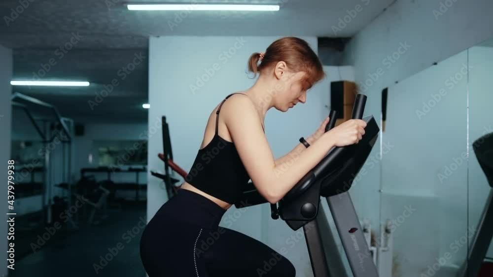 Red-haired girl in black sports uniform in leggings and a top is engaged in gym on simulator in front of mirror and shakes her legs and buttocks. Woman leads healthy lifestyle and does exercise Red-haired girl in black sports uniform in leggings and a top is engaged in gym on simulator in front of mirror and shakes her legs and buttocks. Woman leads healthy lifestyle and does exercise