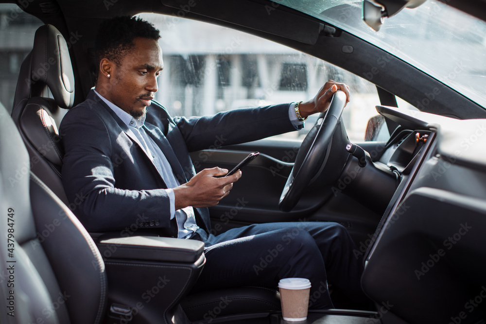 Young african american man in business suit driving car and using modern smartphone. Busy multitasking businessman. Concept of people, transport and concentration.