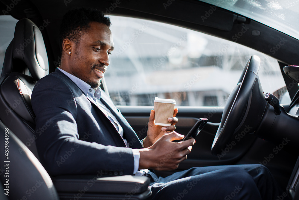 Handsome african man in business suit sitting on driver's seat of his luxury car, drinking coffee and using modern smartphone. Young male taking stop for checking work emails on mobile.