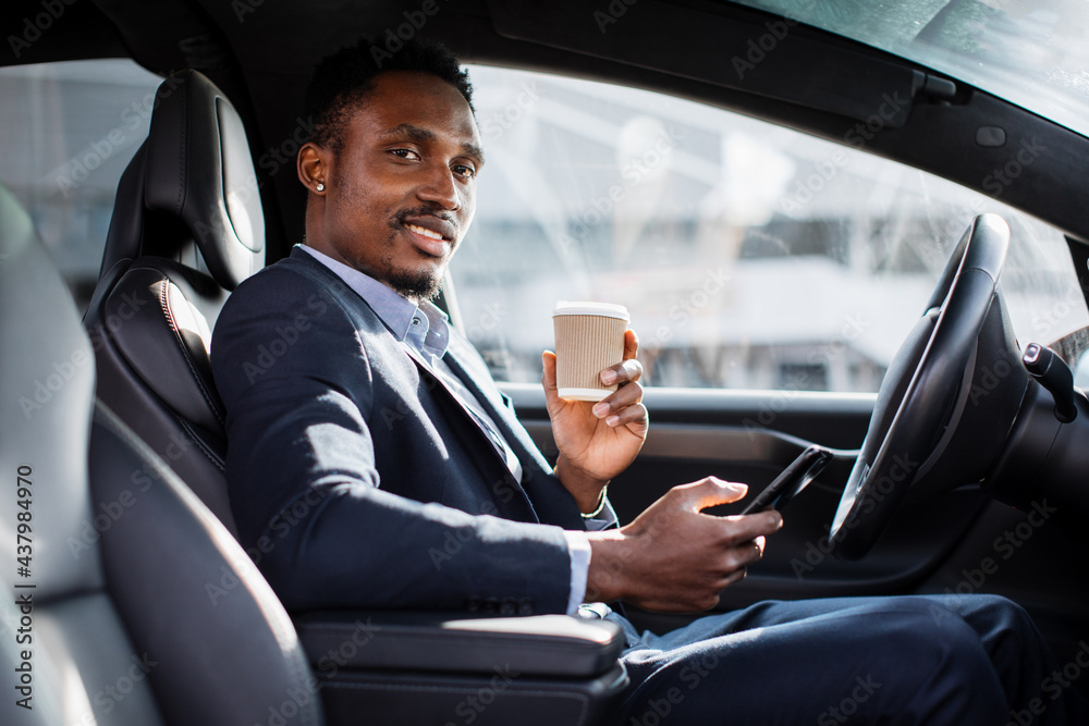 Positive male entrepreneur in business suit having mobile conversation while sitting in car with cup of coffee. African businessman solring working issues on distance. Positive male entrepreneur in business suit having mobile conversation while sitting in car with cup of coffee. African businessman solring working issues on distance.