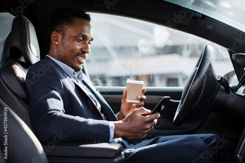 Handsome african man in business suit sitting on driver's seat of his luxury car, drinking coffee and using modern smartphone. Young male taking stop for checking work emails on mobile. © sofiko14