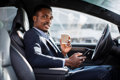 Positive male entrepreneur in business suit having mobile conversation while sitting in car with cup of coffee. African businessman solring working issues on distance. © sofiko14