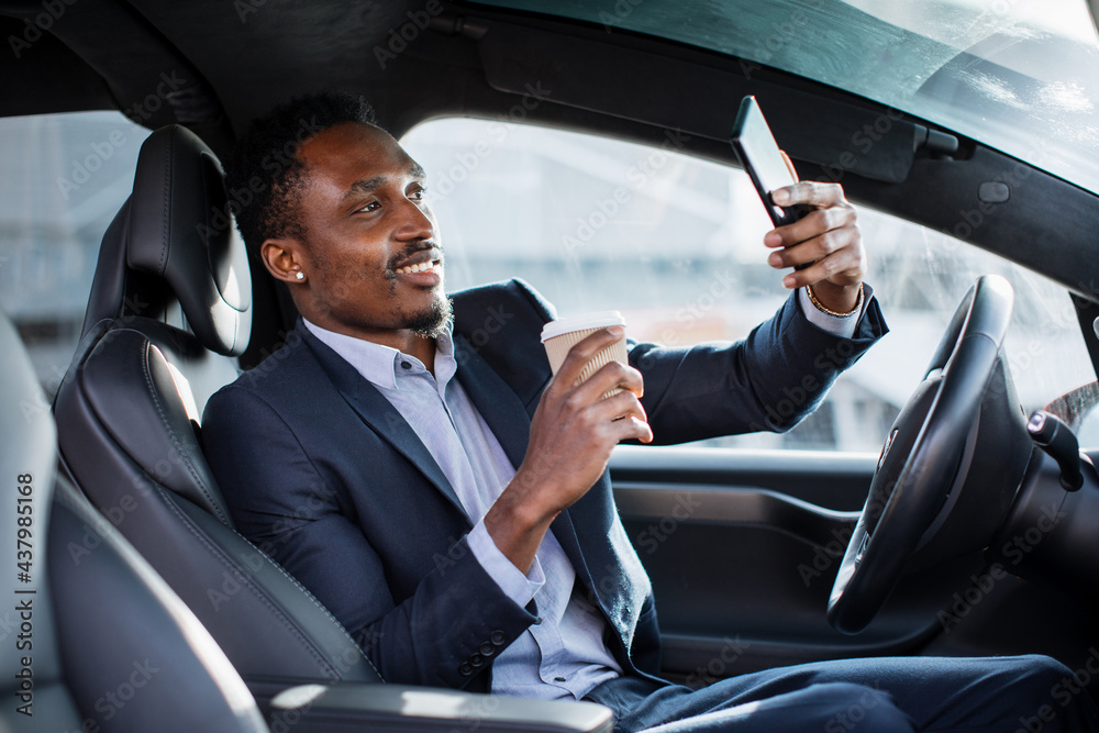 Positive male entrepreneur in business suit having mobile conversation while sitting in car with cup of coffee. African businessman solring working issues on distance.