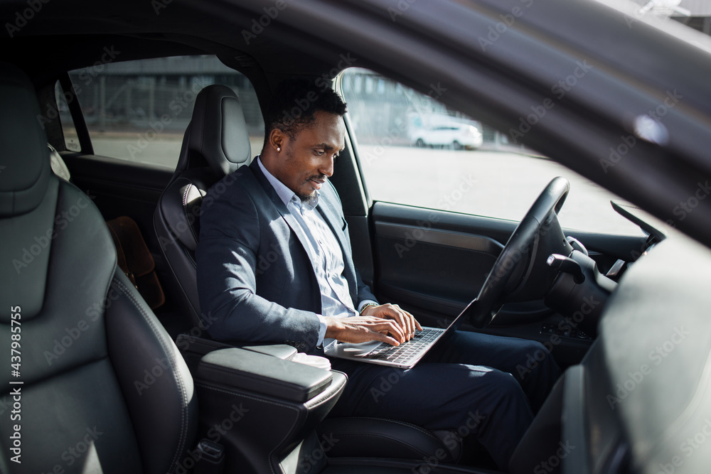 African american man in business suit sitting on driver's seat of modern electric car and typing in wireless laptop. Busy person with portable computer.