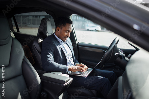 African american man in business suit sitting on driver's seat of modern electric car and typing in wireless laptop. Busy person with portable computer. © sofiko14