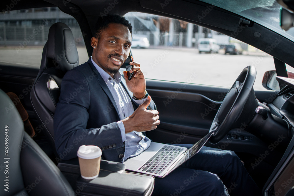 Smiling african man in business suit sitting in modern car with laptop on knees and talking on mobile phone. Young male showing thumb up and looking at camera. Smiling african man in business suit sitting in modern car with laptop on knees and talking on mobile phone. Young male showing thumb up and looking at camera.