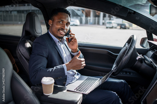 Smiling african man in business suit sitting in modern car with laptop on knees and talking on mobile phone. Young male showing thumb up and looking at camera. © sofiko14