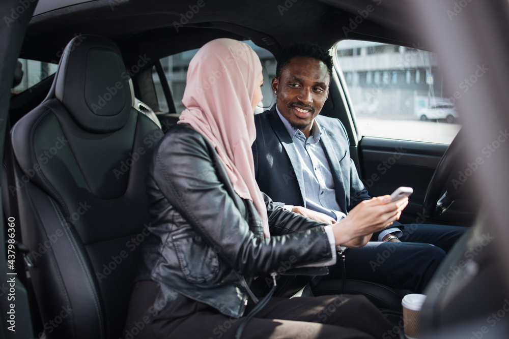 African american businessman in suit and muslim woman in hijab using modern smartphone for remote work while sitting together inside luxury car. Concept of people, gadgets and vehicle.