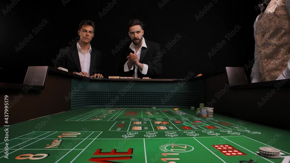 Two men in business suits stand at a green craps table in a casino. The player rolls the dice and wins. Males rejoice with delight and wow emotions. View from inside of game table. Slow motion.