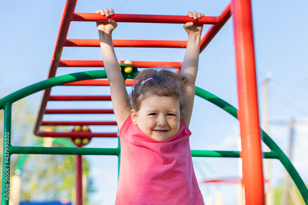 child and sport. little active emotional girl on playground in park, hanging on horizontal bar, doing sports exercise outdoors. happy childhood. Kid play on school or kindergarten yard in summer child and sport. little active emotional girl on playground in park, hanging on horizontal bar, doing sports exercise outdoors. happy childhood. Kid play on school or kindergarten yard in summer