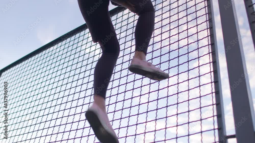 Girl amateur football player climbs over the mesh fence to the sports ground to play football. Soccer training game on court in a sunny day. 4K RAW graded footage.