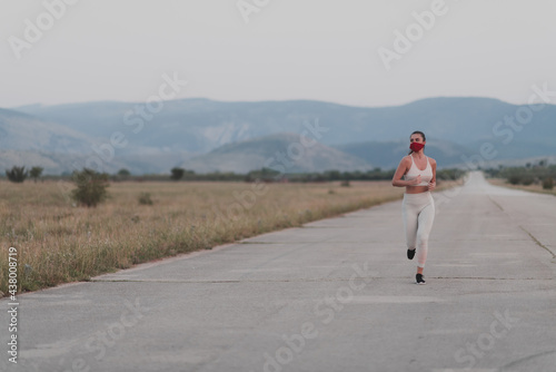 Determined fitness woman in short clothes wearing red protective face mask running outdoors in the city during coronavirus outbreak. Covid 19 and physical jogging activity sport and fitness.