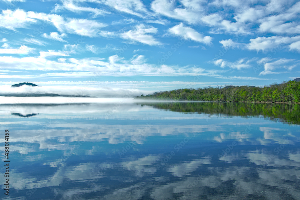 晴れた日の雲の浮かぶ爽快な空と湖畔の森を反射する静水の湖。