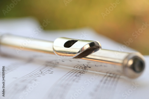Flute, woodwind brass instrument in classical orchestra. Silver modern flute on white sheet music note for education and performance. Song composer on score sheet with green bokeh nature.
