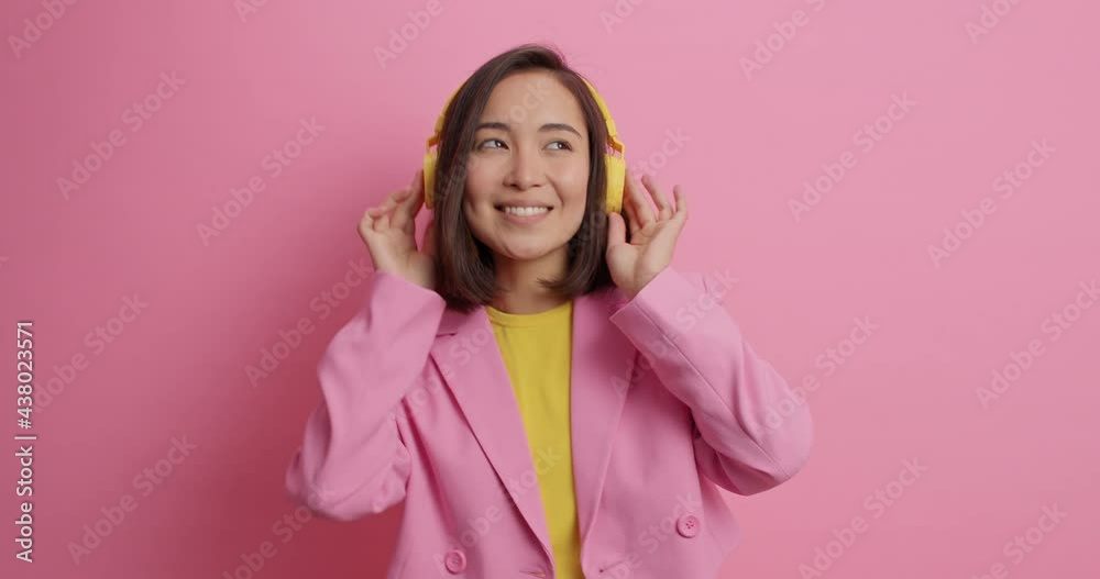 Happy young Asian woman chills indoor catches every bit of music listens favorite playlist keeps hands on headphones wears fashionable clothes isolated over pink background dances to rhythm. Happy young Asian woman chills indoor catches every bit of music listens favorite playlist keeps hands on headphones wears fashionable clothes isolated over pink background dances to rhythm.