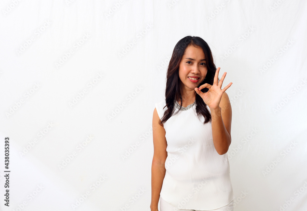 Portrait of a cheerful asian girl dressed in white standing on a white background and doing OK sign with finger and looking at a camera Portrait of a cheerful asian girl dressed in white standing on a white background and doing OK sign with finger and looking at a camera
