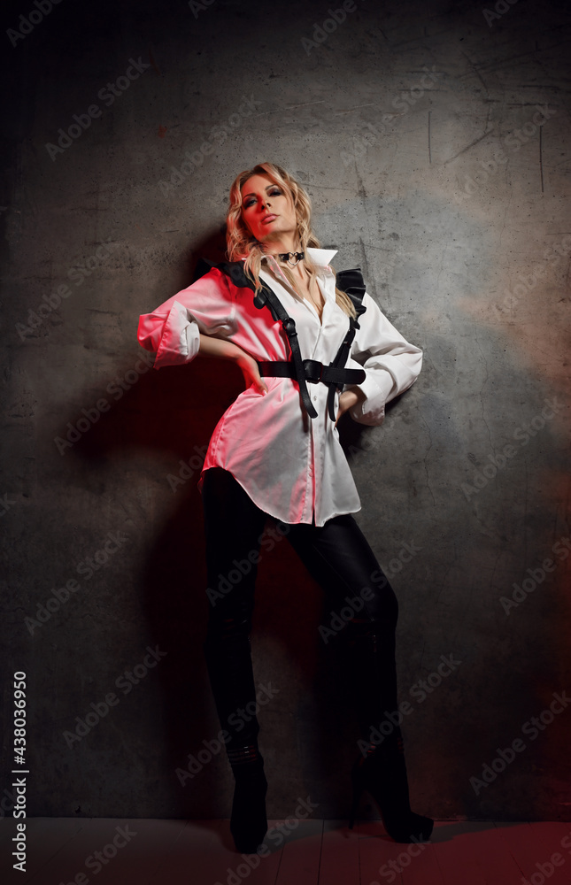 Insolent blonde woman mistress in leather pants, white shirt with leather  belts and cap stands at concrete wall looking down at camera. Fashion,  vogue, sexy stylish look for woman concept Stock Photo