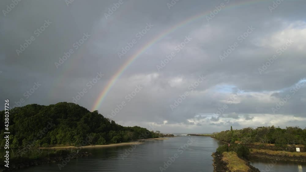 Travelling along a scenic river towards the open ocean with a large colorful rainbow and cloudy sky in the distance. Moving drone view Travelling along a scenic river towards the open ocean with a large colorful rainbow and cloudy sky in the distance. Moving drone view