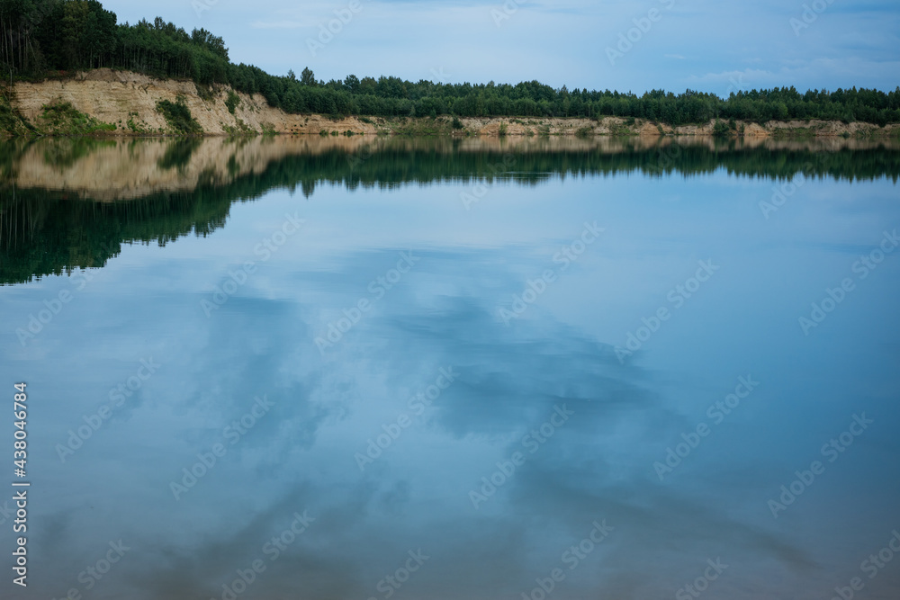 azure, water landscape against the blue sky. spruce forest, sandy cliff and clouds are reflected in the water surface.