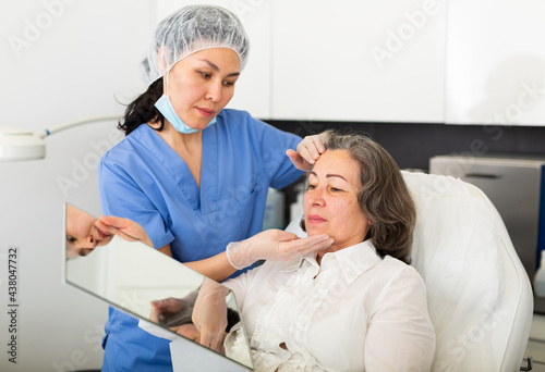 Asian female cosmetologist giving professional advice to elderly woman before hardware facial skin treatments in cosmetological office .