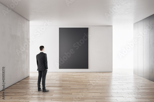 Businessman in modern concrete interior with empty poster looking at mockup place for your advertisement.