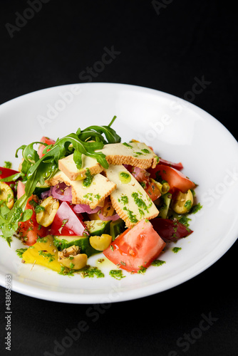 Vegetable salad with cheese, fresh romatoes and cucumbers, green olives served in a white plate over black background.