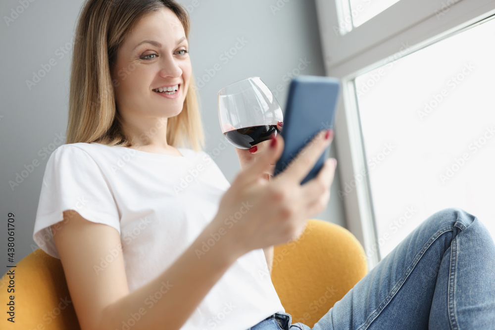 Young woman holding mobile phone in her hands and drinking red wine from glass at home Young woman holding mobile phone in her hands and drinking red wine from glass at home