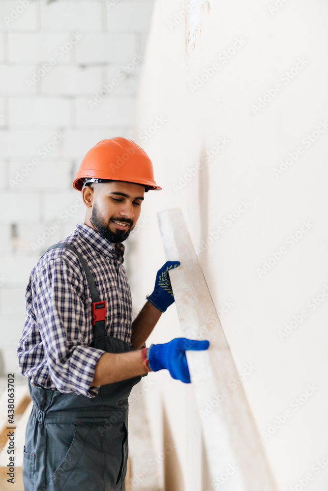A worker in overall and cap, holding a comb putty-knife and covering a wall surface with adhesive paste before laying decorative stone. Facing work, renovation and interior design concept A worker in overall and cap, holding a comb putty-knife and covering a wall surface with adhesive paste before laying decorative stone. Facing work, renovation and interior design concept