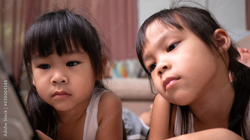 Two sibling girls lying on the sofa and watching digital tablets together in the living room. Two sibling girls lying on the sofa and watching digital tablets together in the living room.
