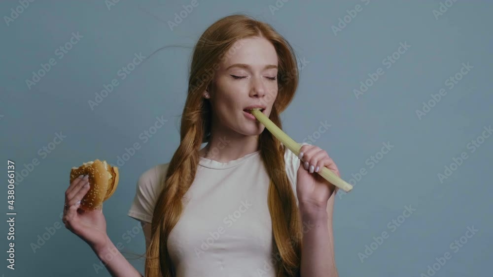 Ginger woman with red hair and freckles at studio biting a leek while holding a hamburger Ginger woman with red hair and freckles at studio biting a leek while holding a hamburger