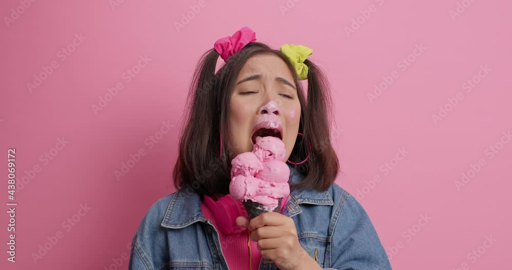 Dejected Asian woman sobs and cries has troubles in life licks ice cream tries to sooth herself smeared with sweet dessert wears denim jacket isolated over pink background. Upset teenager with gelato Dejected Asian woman sobs and cries has troubles in life licks ice cream tries to sooth herself smeared with sweet dessert wears denim jacket isolated over pink background. Upset teenager with gelato
