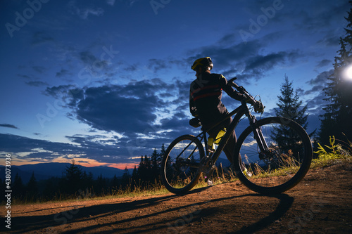 Back view of young man sitting on bicycle under beautiful night sky. Male bicyclist resting on hillside under blue cloudy sky while riding bicycle. Concept of sport, biking and active leisure.