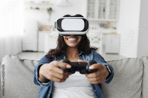 Happy african american woman in vr glasses with joystick, passionately playing in online games sitting on sofa © Prostock-studio