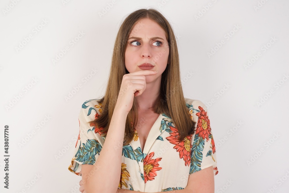 Shot of contemplative thoughtful Young woman keeps hand under chin, looks thoughtfully upwards, dressed in casual clothes, poses over white background with free space for your text