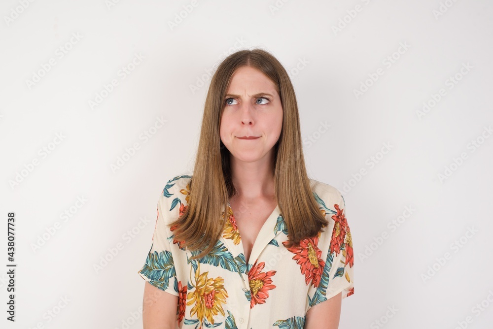Photo of amazed puzzled young Caucasian female with ginger hair knot, curves lips and has worried look, sees something awful in front, isolated on white background, dressed in jean overalls.