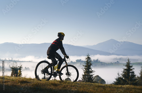 Silhouette of man cyclist in cycling suit riding bike on grassy hill. Male bicyclist enjoying the view of majestic mountains during bicycle ride. Concept of sport, bicycling and nature.