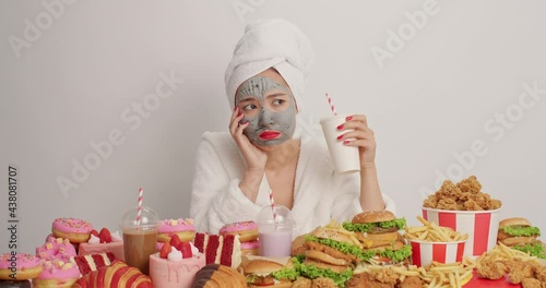 Bored lonely depressed Asian woman drinks soda feels unhappy applies beauty mask on face tries to sooth herself with delicious fast food affords eating harmful snack during domestic pajama party.