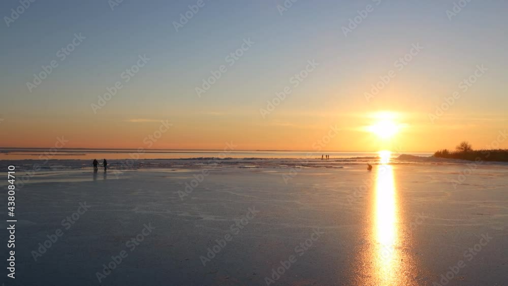 The frozen lake in winter on sunset. Children and adults have fun walking and rolling on ice-covered water. The sun shines brightly The frozen lake in winter on sunset. Children and adults have fun walking and rolling on ice-covered water. The sun shines brightly