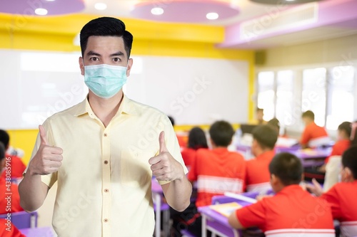 Portrait handsome young asian man wearing mask protection from covid 19 and thumbs up sign on blurred student study in class at school. Education concept.