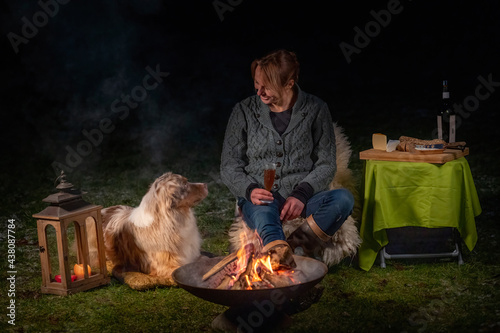Young woman drinks red wine by the campfire in the forest. Her Australian Shepperd is lying next to her on a rug in the grass. The dog looks at its owner. Enjoying food and drink in leisure time in