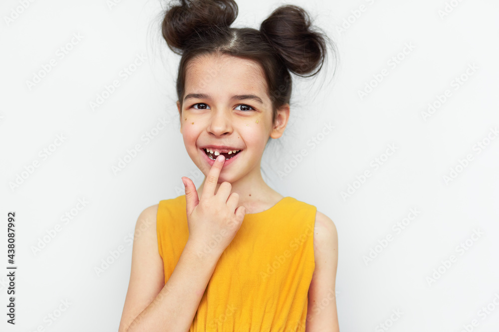 A cute little girl in yellow dress, funny hairstyle, showing empty space with growing first permanent molar, isolated on grey background. A child has lost the first milk tooth. Kid missing baby tooth. A cute little girl in yellow dress, funny hairstyle, showing empty space with growing first permanent molar, isolated on grey background. A child has lost the first milk tooth. Kid missing baby tooth.